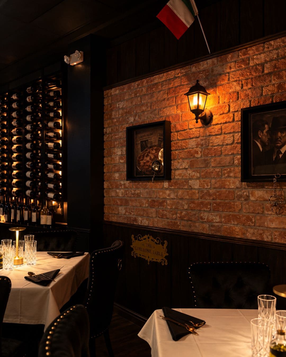 Brick wall with a brass sconce and framed portraits beside the lit wine wall, plaque reading The Corleone Table