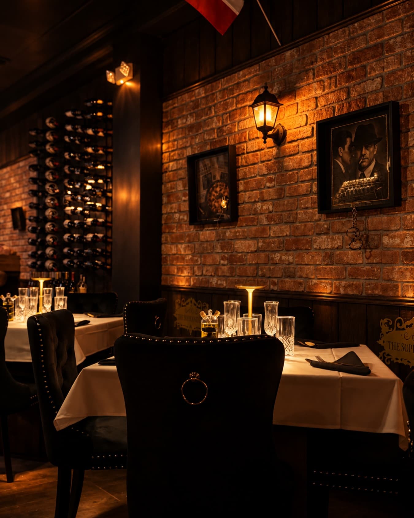 Tables laid with cut crystal and brass lamps under a lantern on the brick wall, framed portraits above, the lit wine rack behind and the Soprano plaque on the wainscot