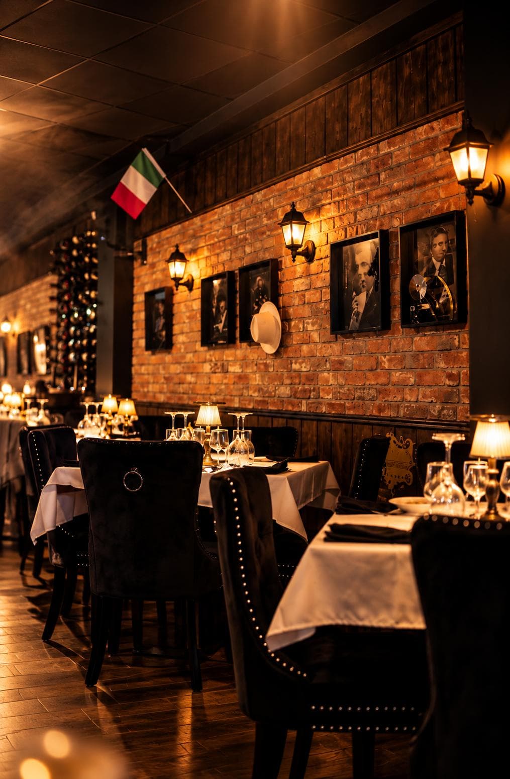The dining room along the brick wall: brass lanterns, framed black-and-white portraits, a white fedora and an Italian flag above tables laid in white