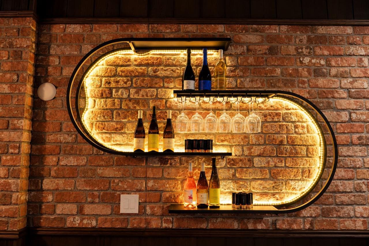 A rounded shelf lit from behind on the brick wall, with white, rosé and red bottles and a row of hanging wine glasses