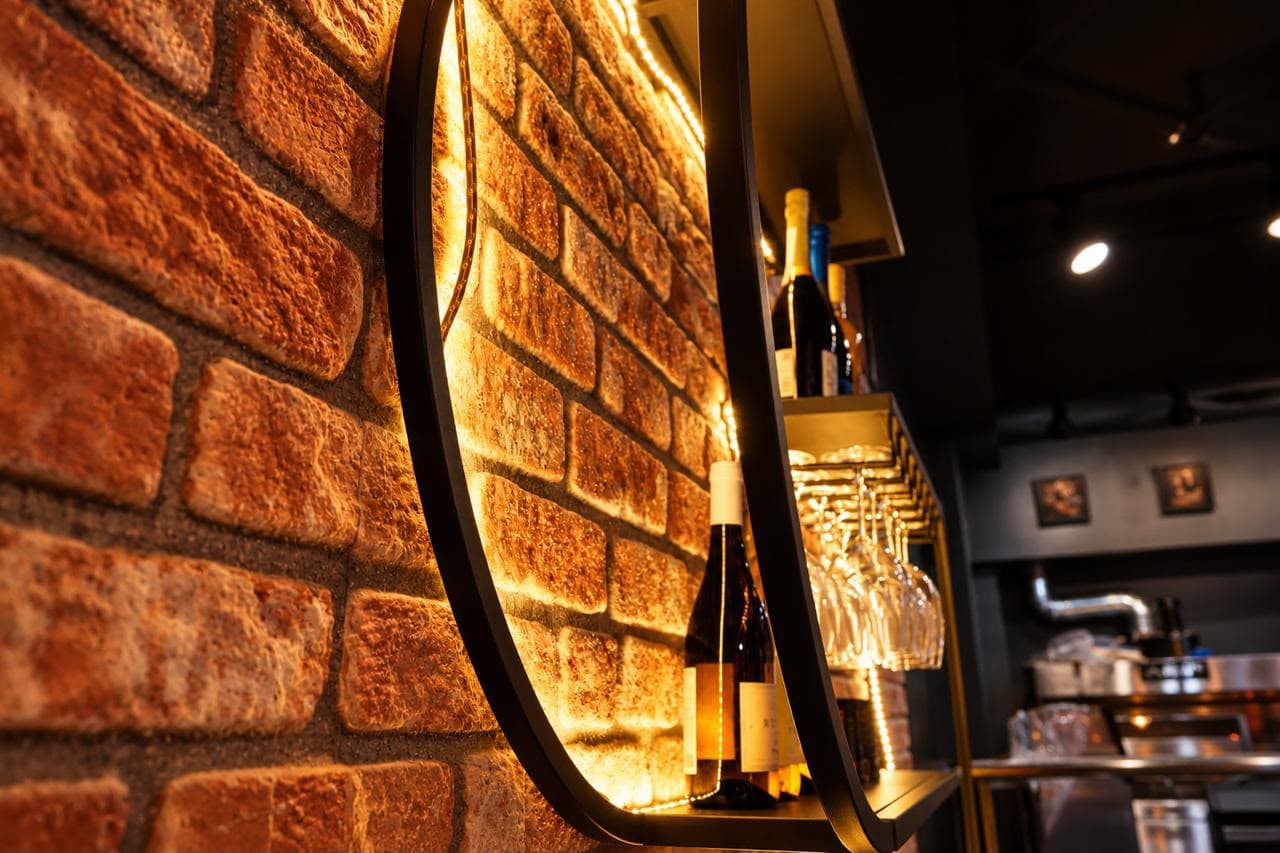A curved shelf lit from behind on the brick wall, seen from the side: a bottle, hanging wine glasses and the bar out of focus beyond