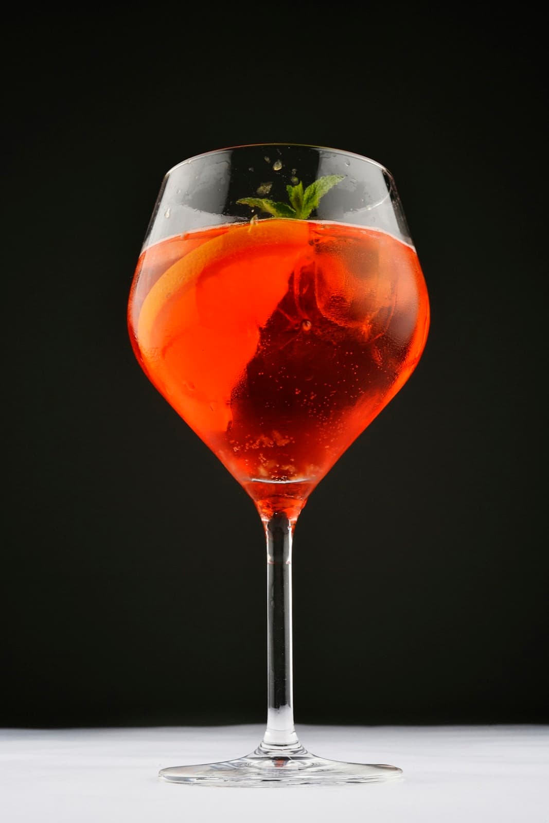 An Italian spritz in a stemmed glass with an orange slice and mint, on a dark background